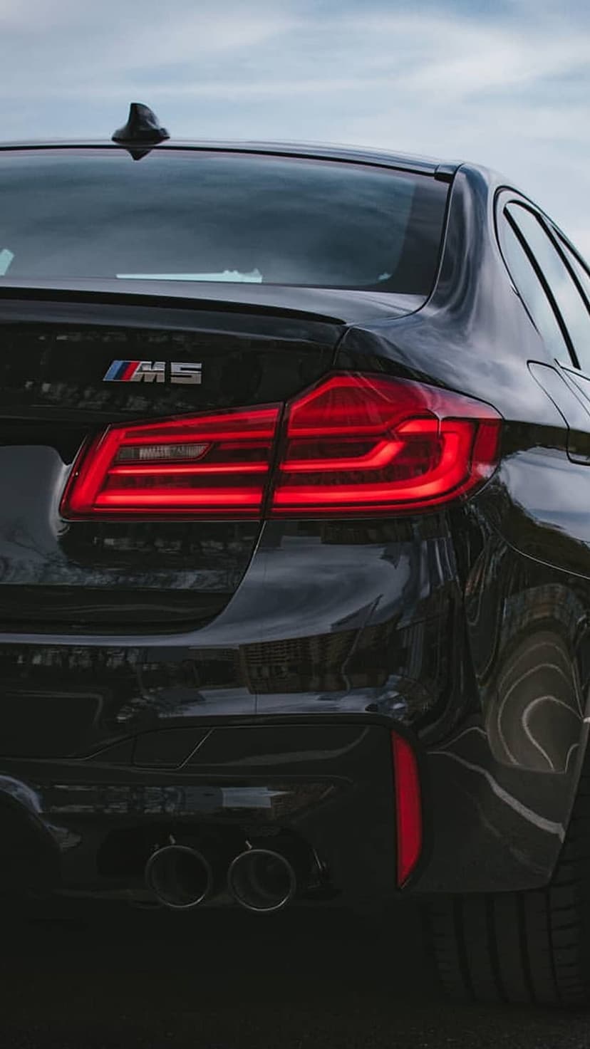BMW M5 rear bumper with glowing red LED taillights, quad exhaust pipes, and a glossy black paint finish.