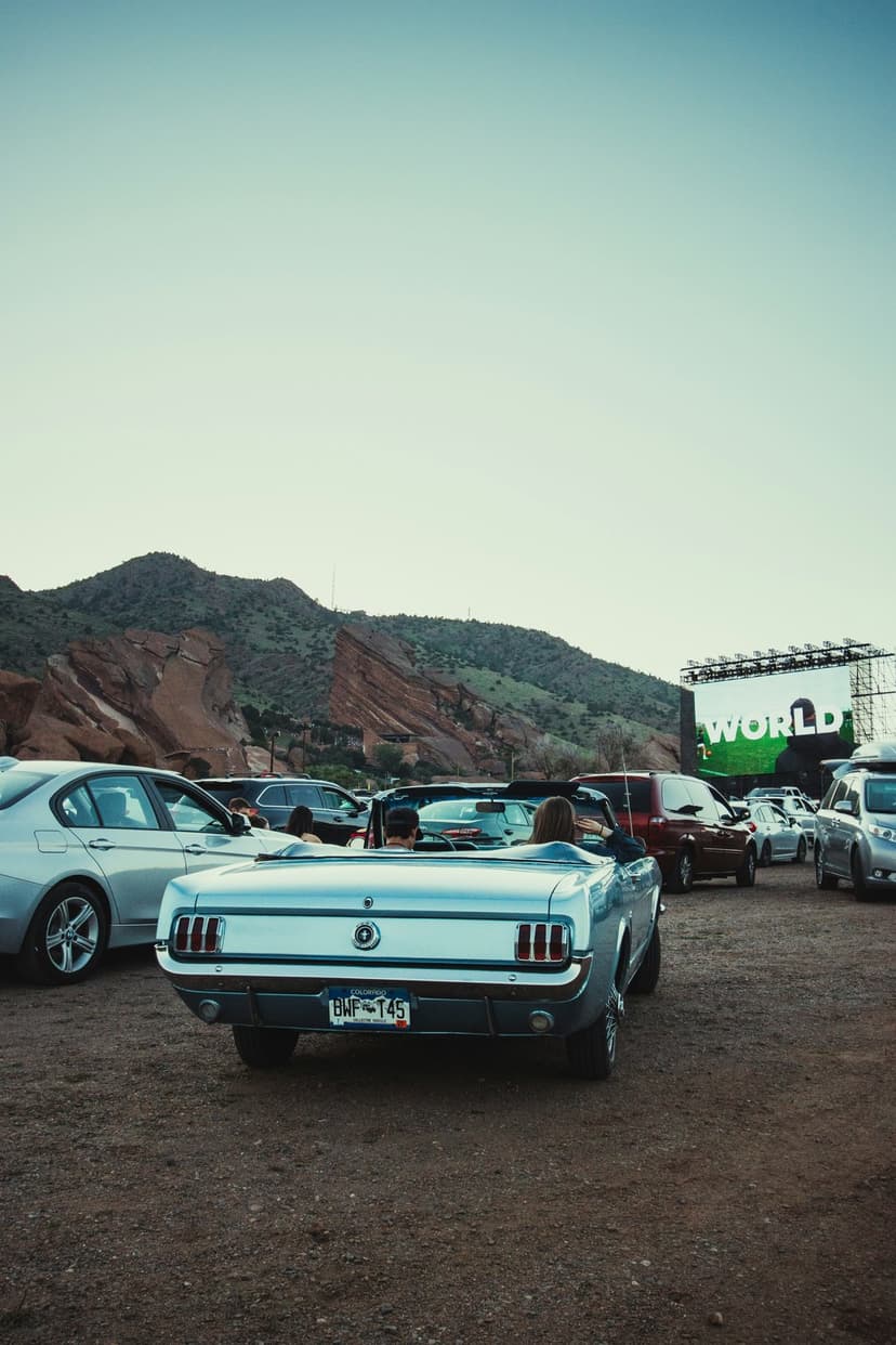 Classic Mustang convertible at a drive-in cinema with red rock mountains under a soft, pale evening sky.