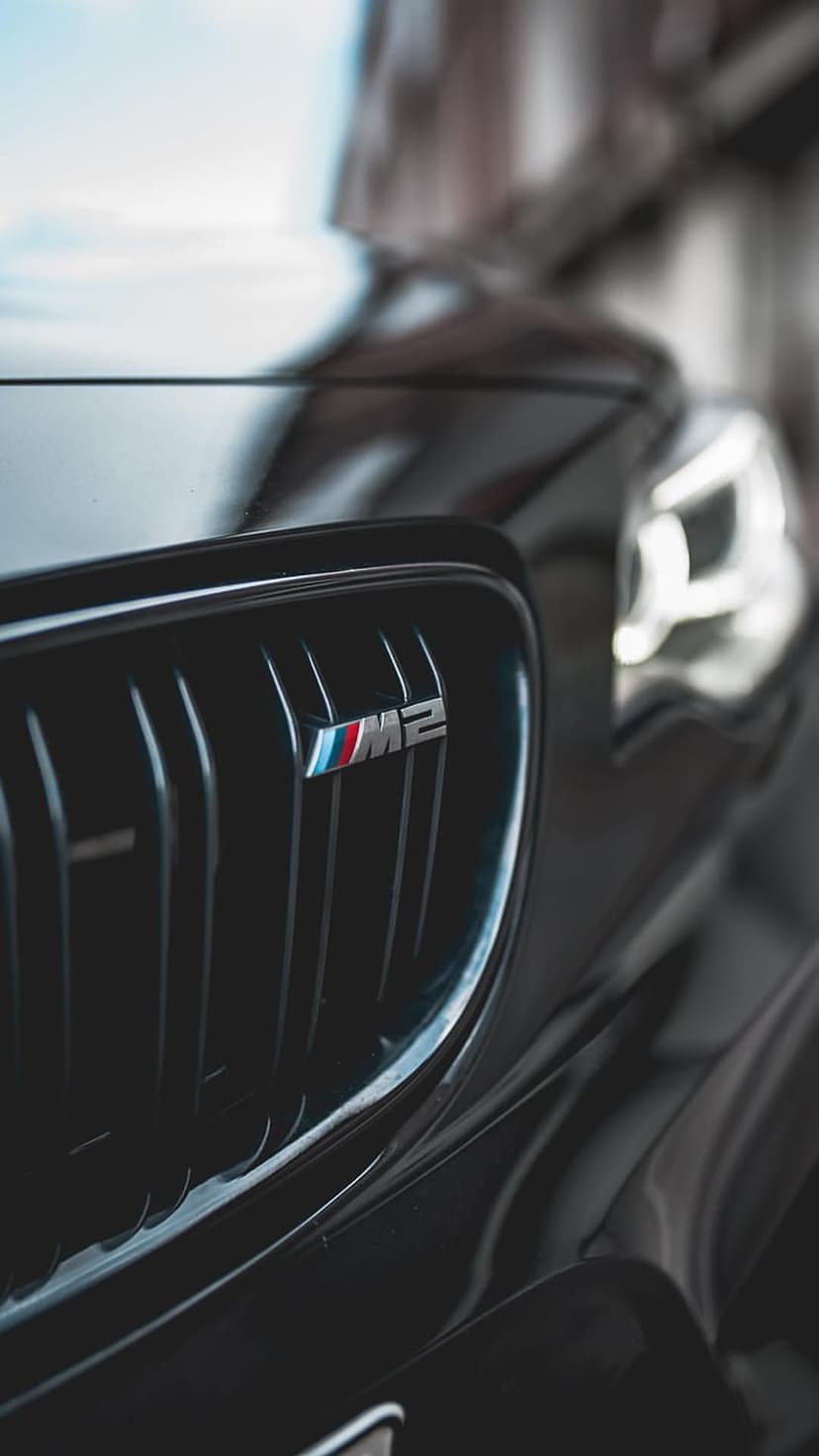 BMW M2 badge on a black kidney grille next to a sharp LED headlight with blue accents and dark reflections.