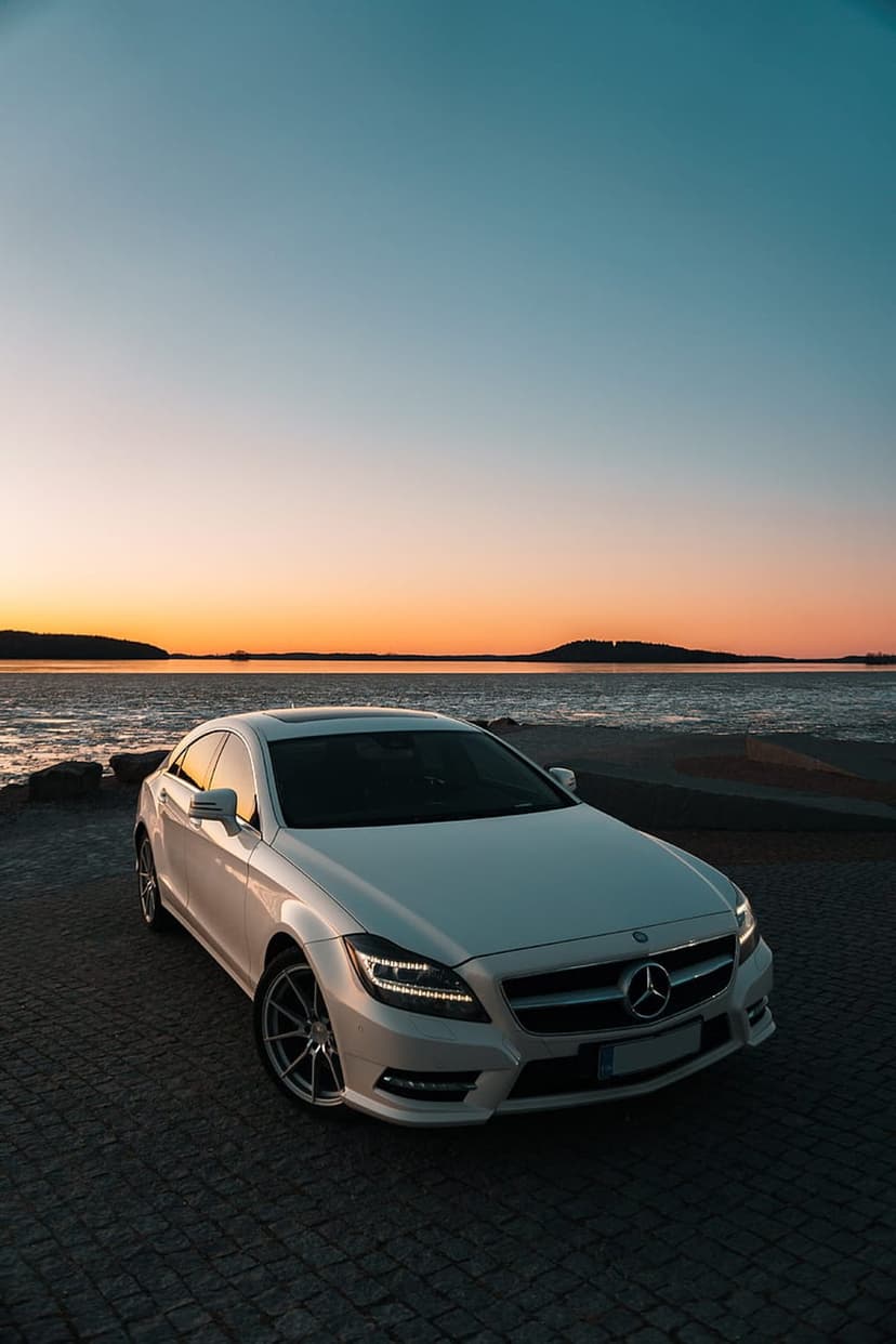 White Mercedes CLS sedan parked on cobblestones by the sea during a golden sunset with distant islands.