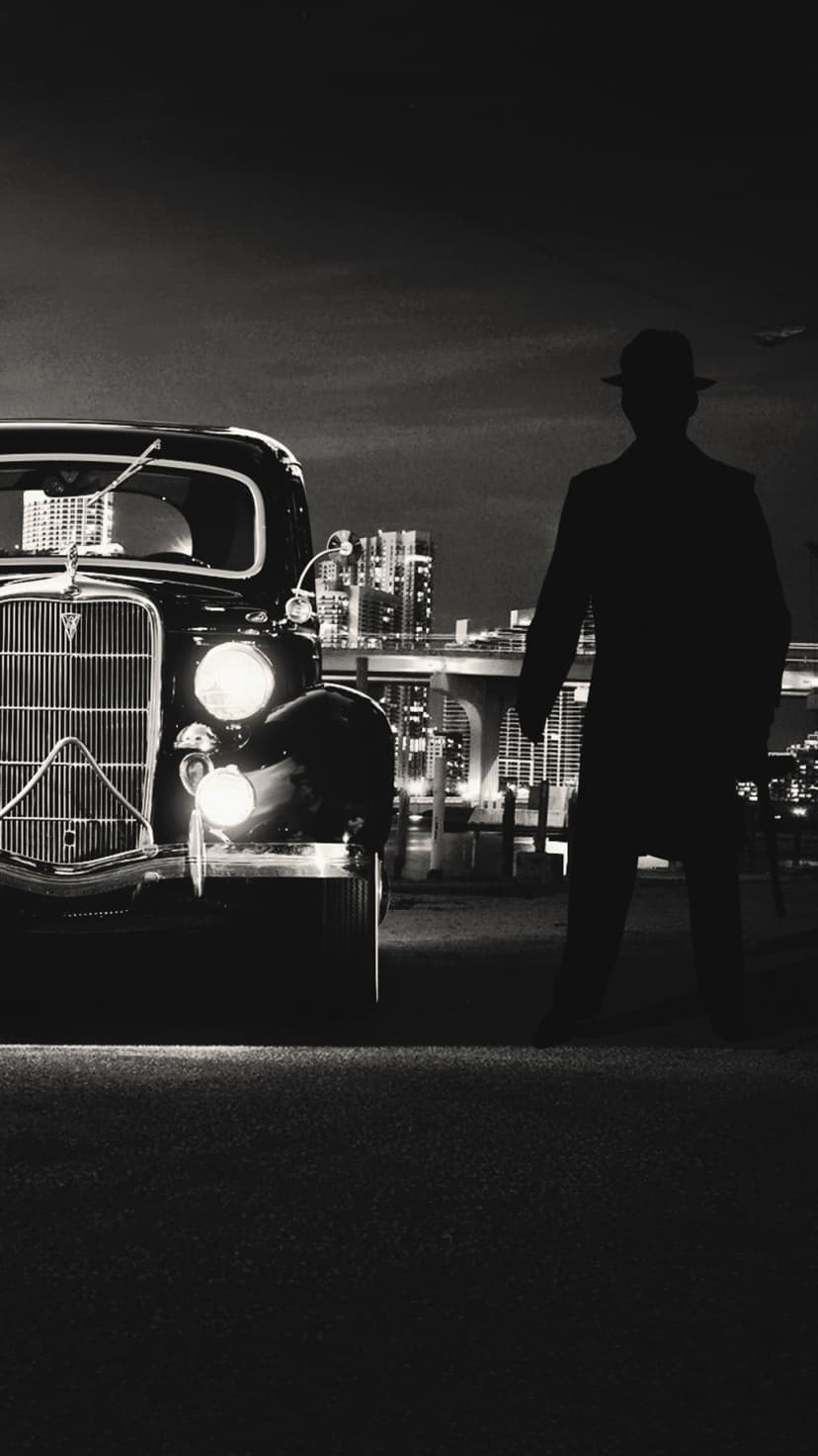 Silhouette of a man in a fedora standing by a vintage car with glowing headlights against a city skyline.