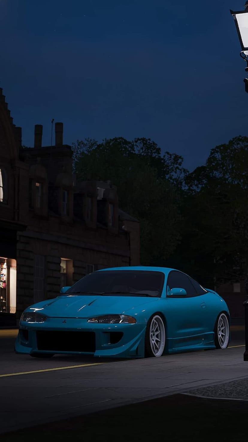Blue Mitsubishi Eclipse with white rims parked on a dark cobblestone city street under glowing night streetlights.