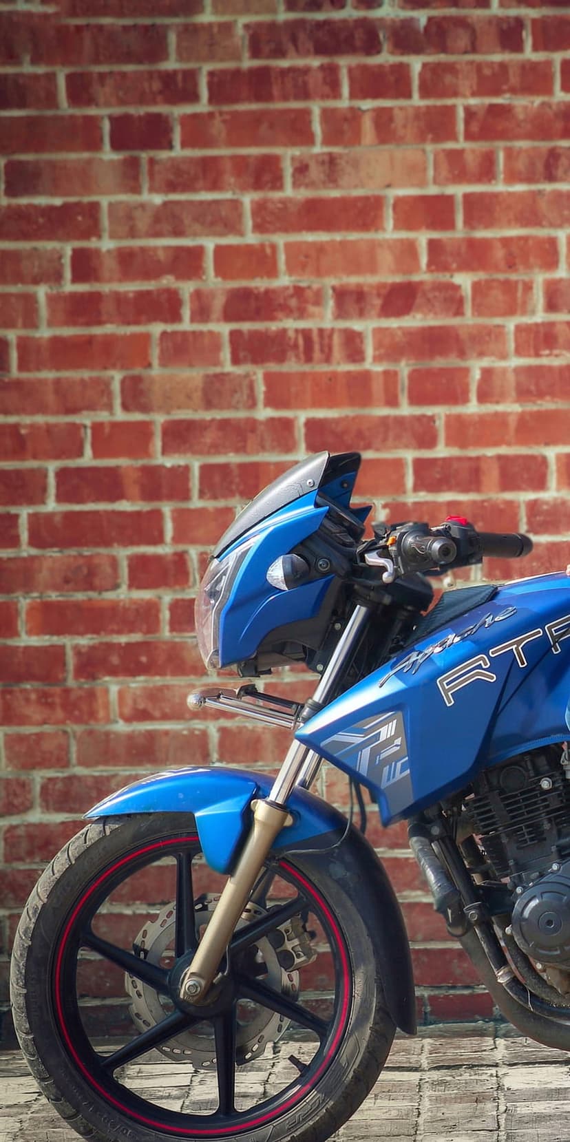 Blue Apache motorcycle with a red-rimmed front wheel leans against a rustic textured brick wall in daylight.