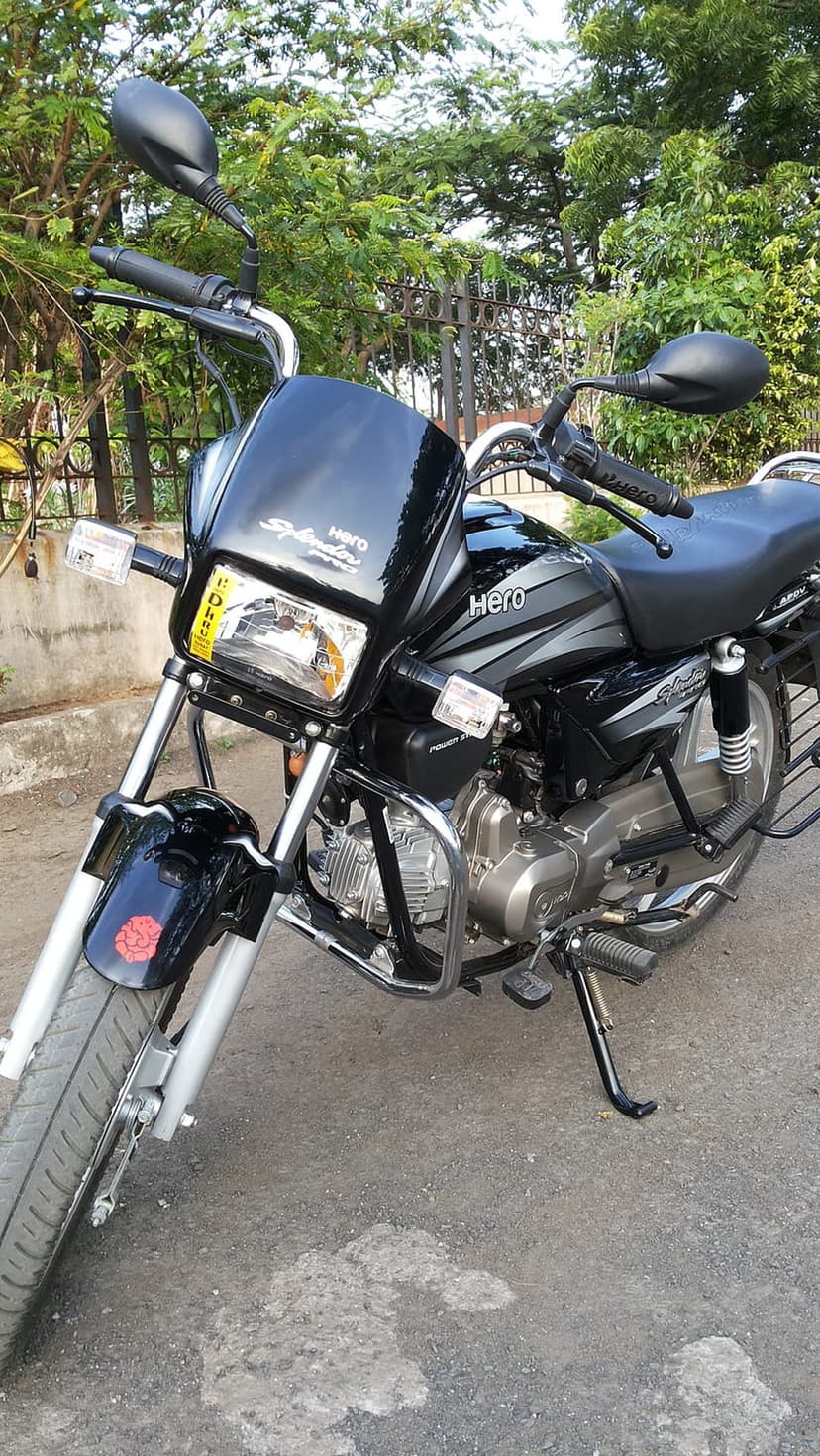 Black Hero Splendor motorcycle parked on an asphalt road with a chrome engine guard and green forest background.