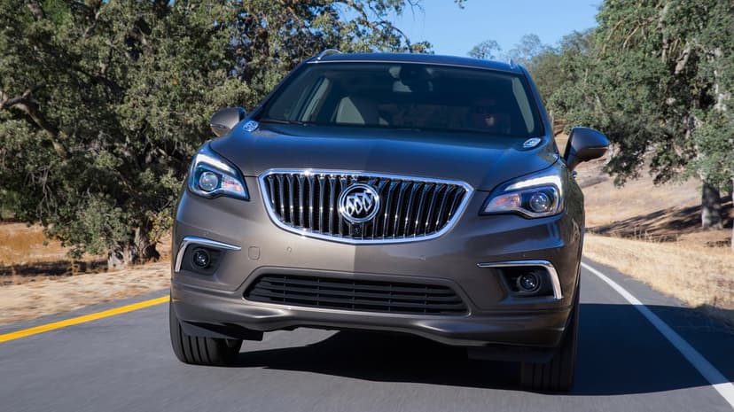 Buick Envision SUV driving on a sunlit road with green trees and golden hills in the background.