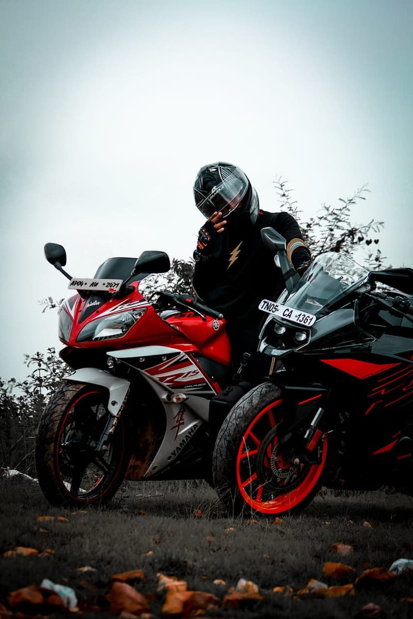 Motorcyclist in a helmet stands beside red and black Yamaha R15 sportbikes under a cloudy sky in a dry field.