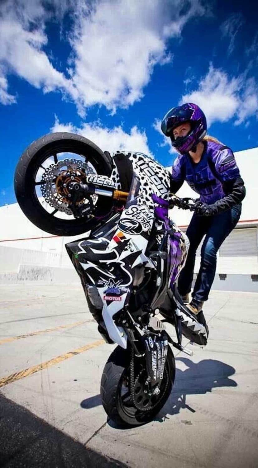 Female motorcyclist in a purple helmet performs a wheelie on a sportbike under a bright blue sky with white clouds.
