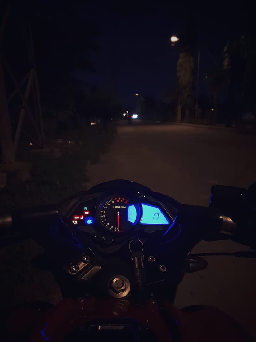 Motorcycle instrument cluster glowing blue at night with a needle tachometer and blurred streetlights behind.