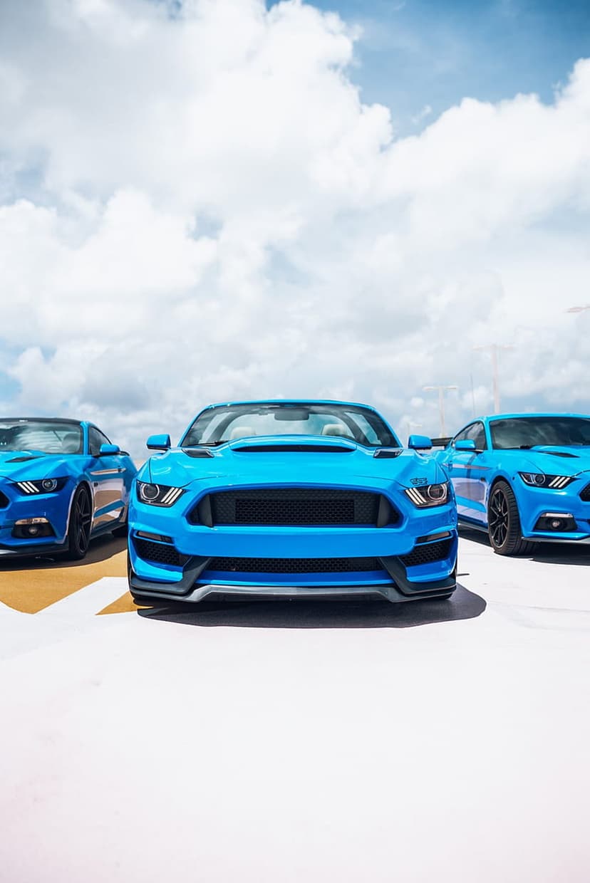 Three blue Ford Mustangs parked on yellow and white pavement under a bright sky with fluffy white clouds.
