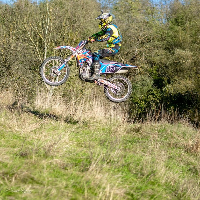 Motocross rider on a red and white bike jumping over a sunlit grassy hill with trees in the distant background.