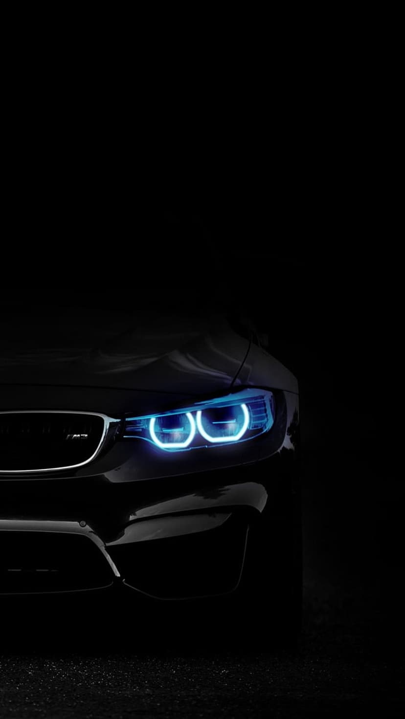BMW car headlights with blue glowing angel eye rings emerge from deep shadows in a close-up night shot.