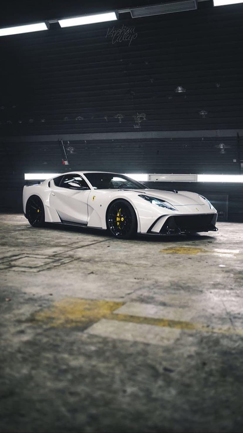White Ferrari supercar with yellow calipers parked on a textured concrete floor in a dimly lit industrial garage.