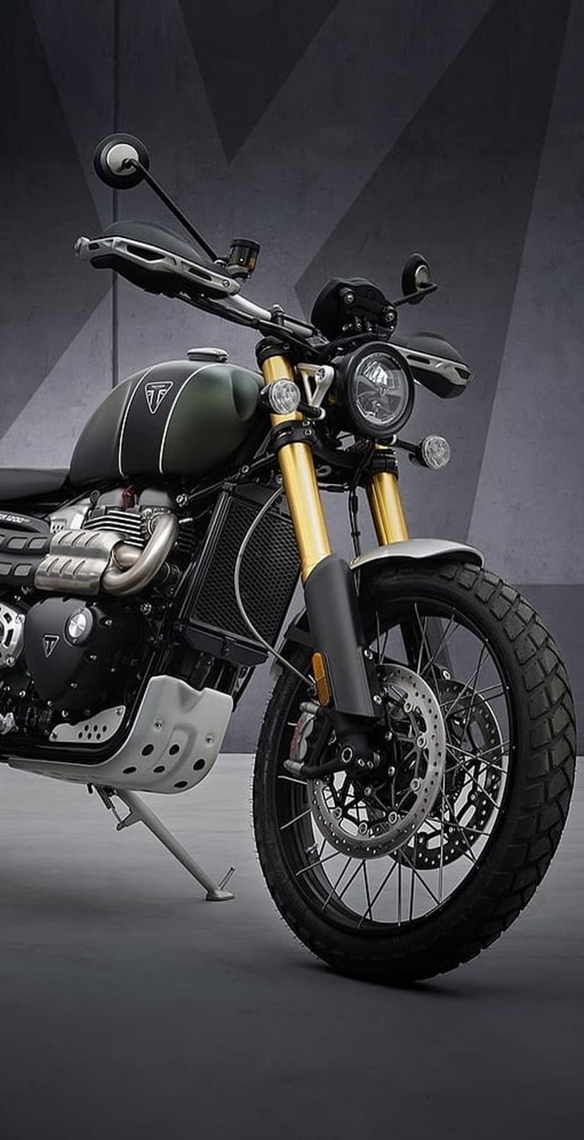 Triumph Scrambler motorcycle featuring gold front forks, knobby tires, and a green fuel tank on a grey surface.