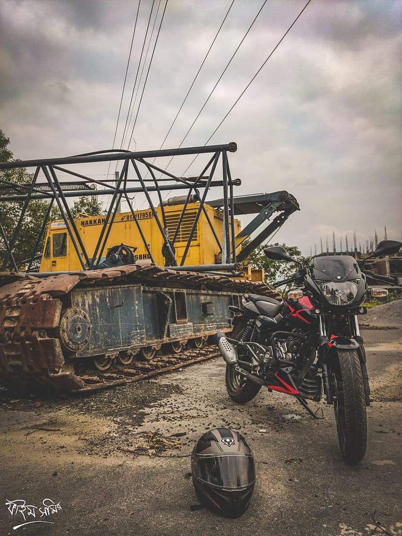Black and red motorcycle parked beside a towering rusty crane at an overcast urban construction site.