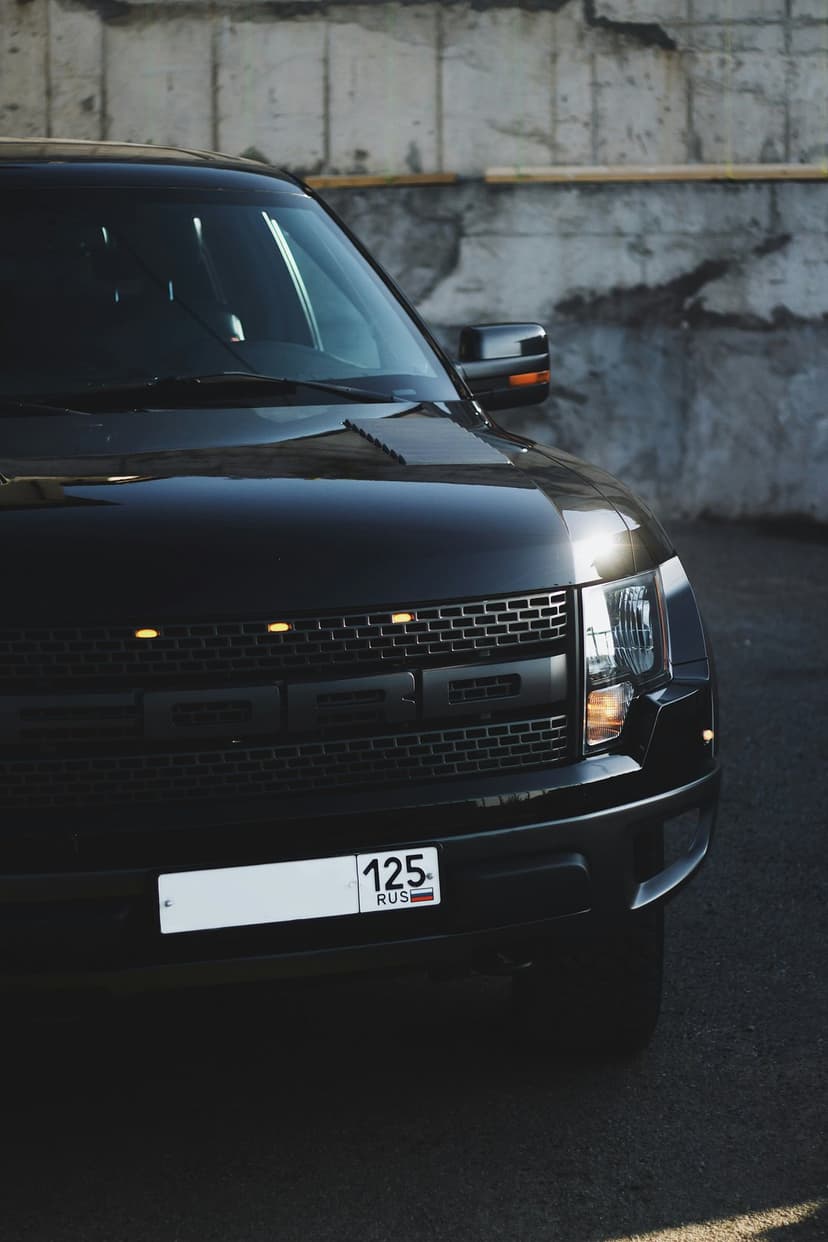 Ford F-150 Raptor truck front grille with amber lights and a license plate against a textured concrete wall.