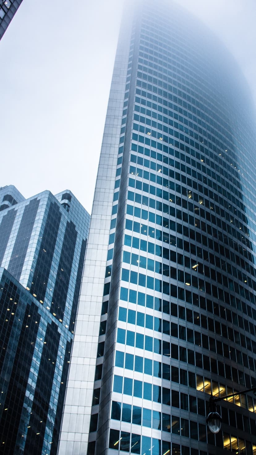 Modern glass skyscrapers rise into a dense layer of white fog from a steep, dramatic upward camera perspective.