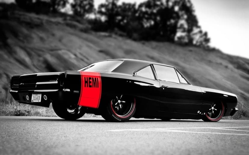 Black muscle car with a red Hemi stripe on a road against a blurred monochrome hillside background.