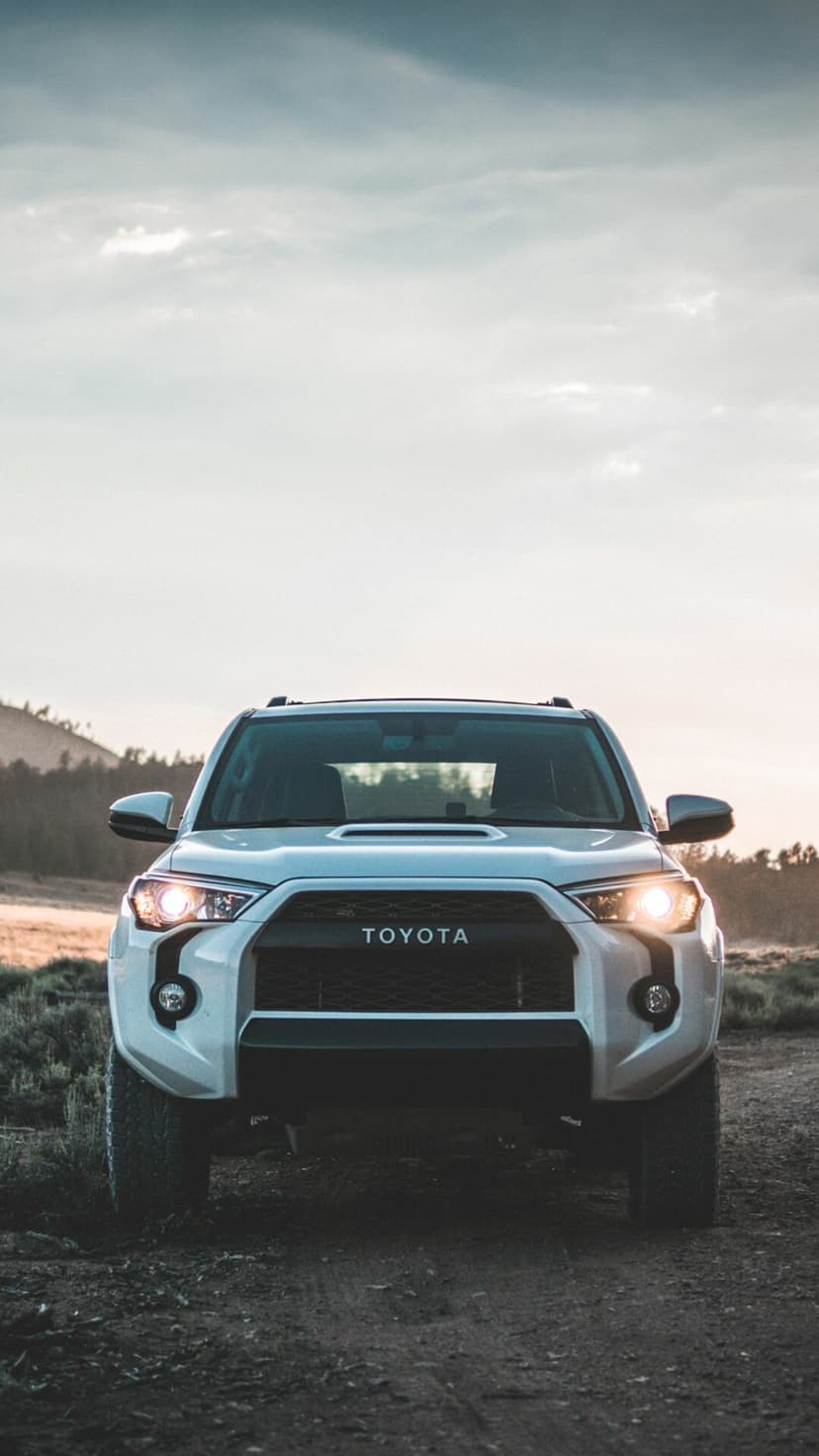 Front view of a white Toyota 4Runner with headlights on, parked on a dirt trail at dawn with mountains behind.