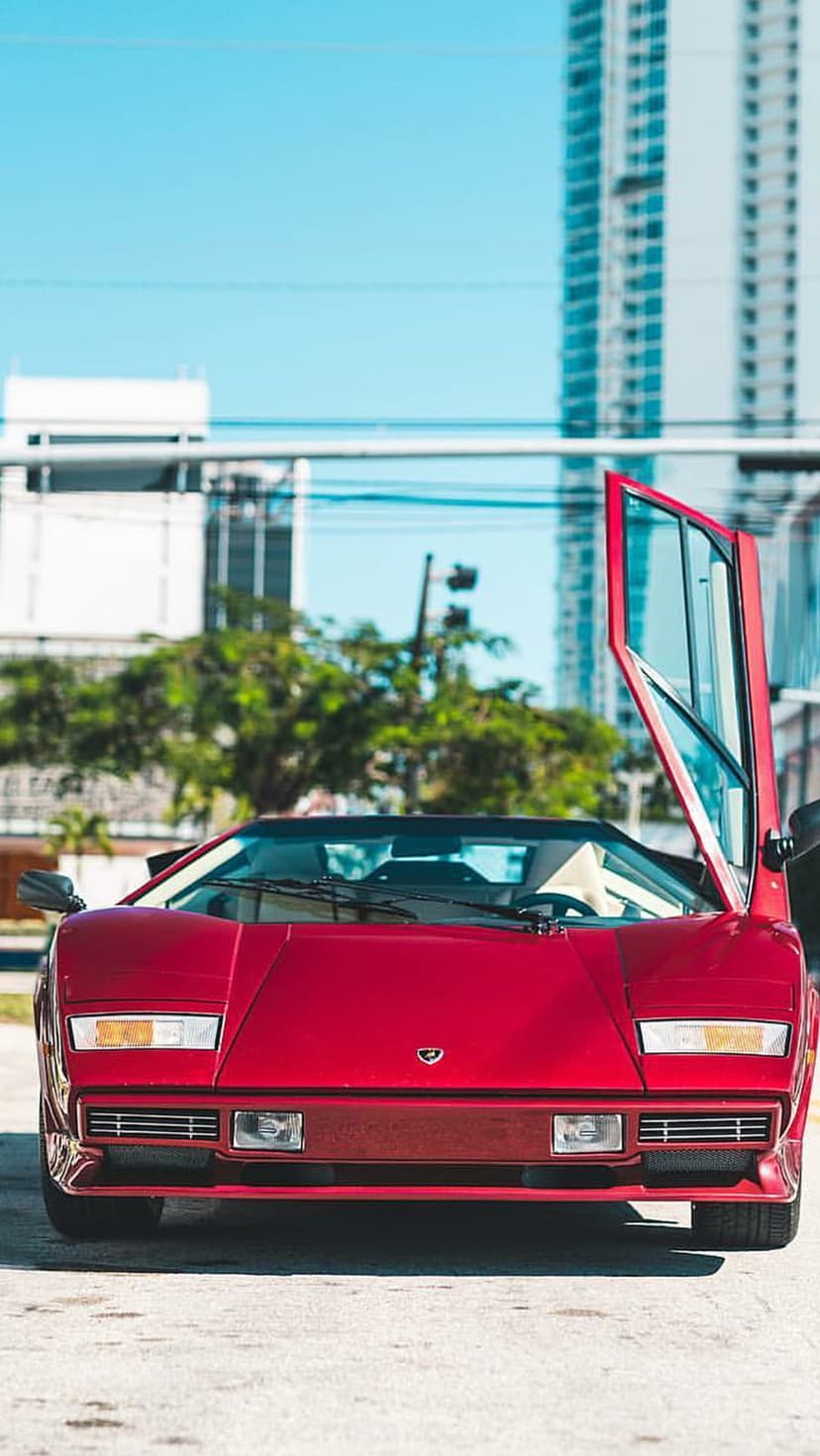 Red Lamborghini Countach with open scissor doors parked on a city street under a blue sky and modern buildings.