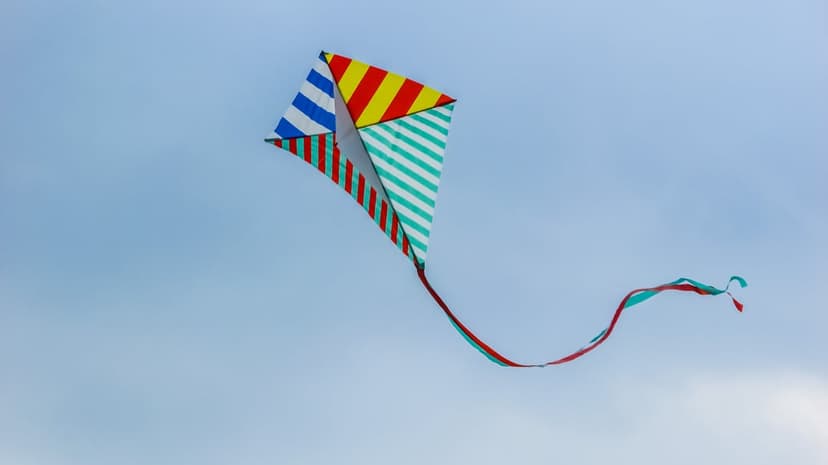 Multicolored striped diamond kite with red and turquoise ribbon tails soaring through a clear pale blue sky.