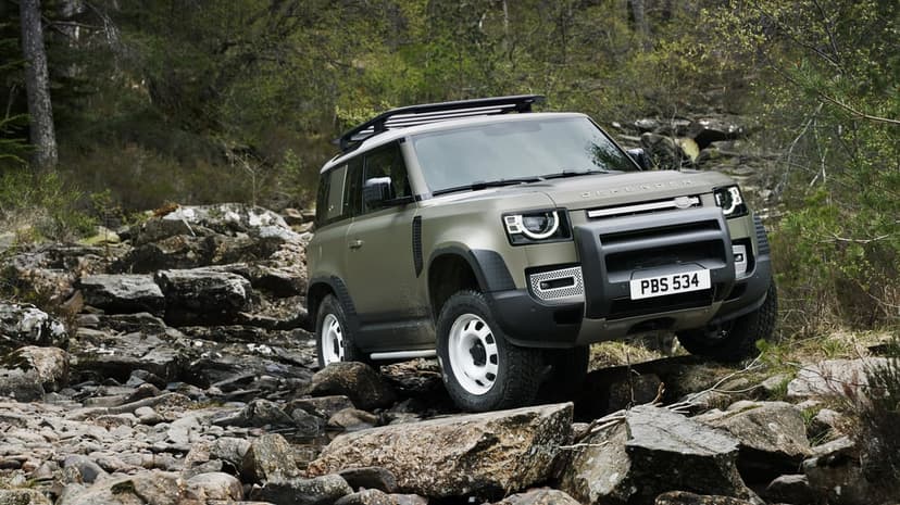 Land Rover Defender 90 off-roading over grey rocks in a green forest with white steel wheels and roof rack.