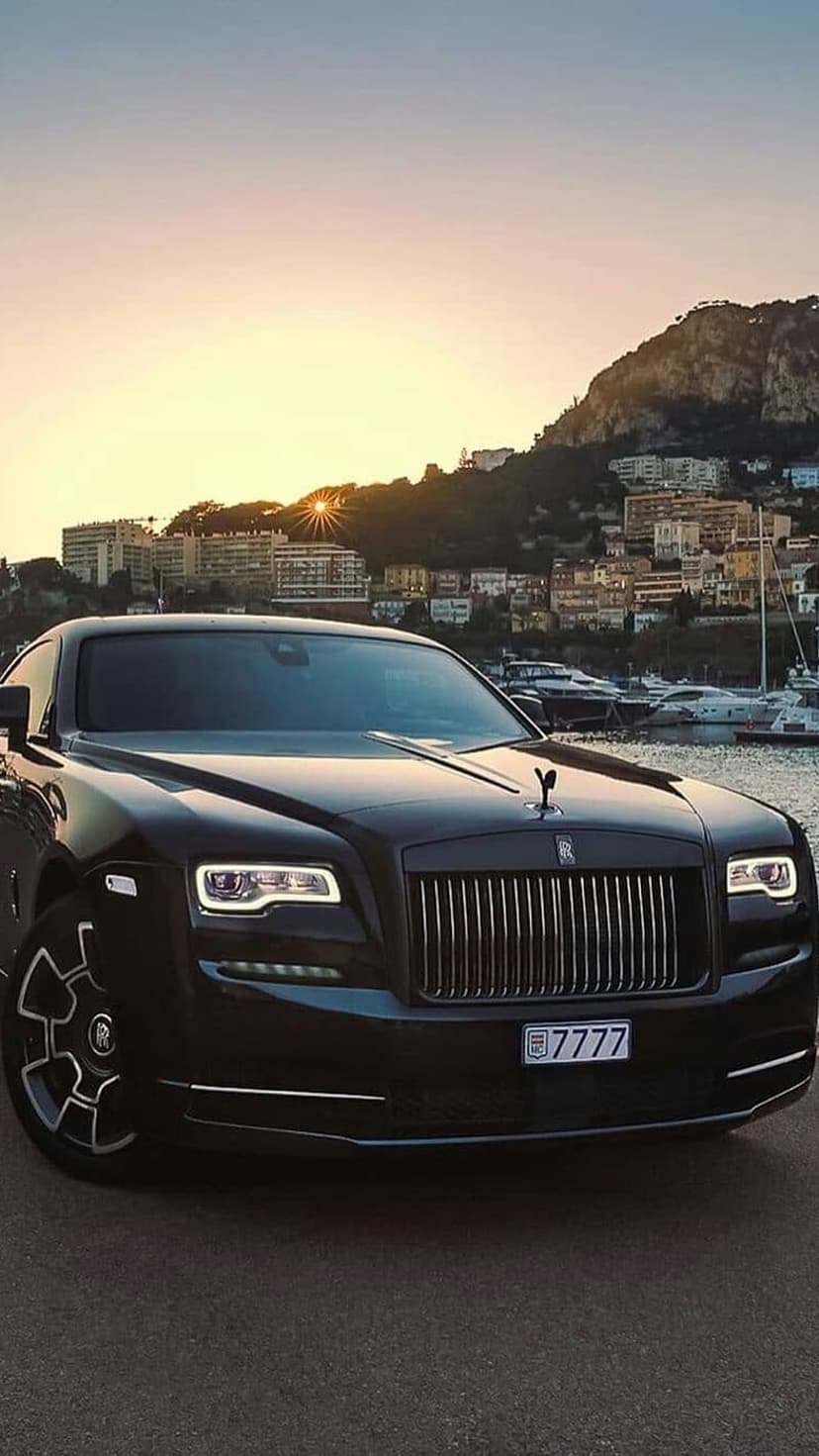 Black Rolls-Royce Wraith parked near a coastal harbor at sunset with city lights and yachts in the background.