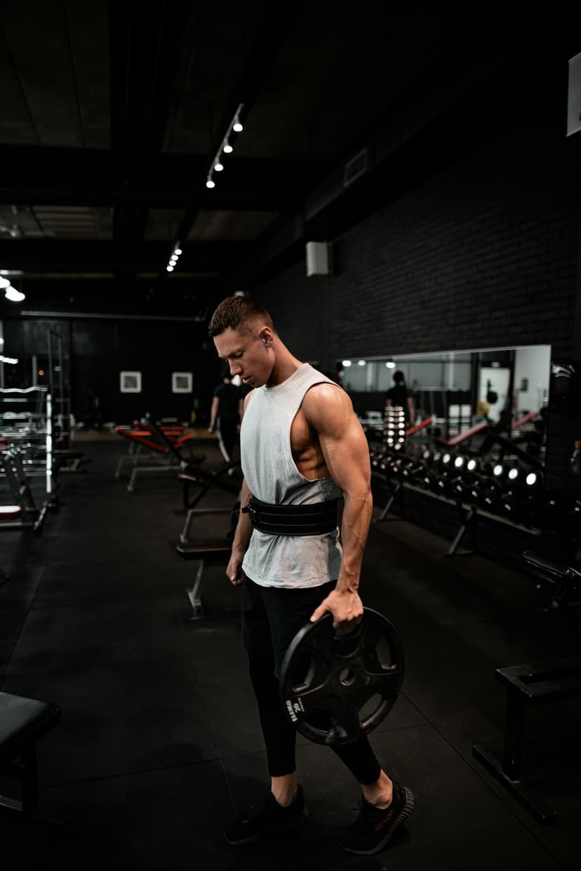 Muscular man in tank top holds a heavy iron weight plate while standing inside a dimly lit fitness center.