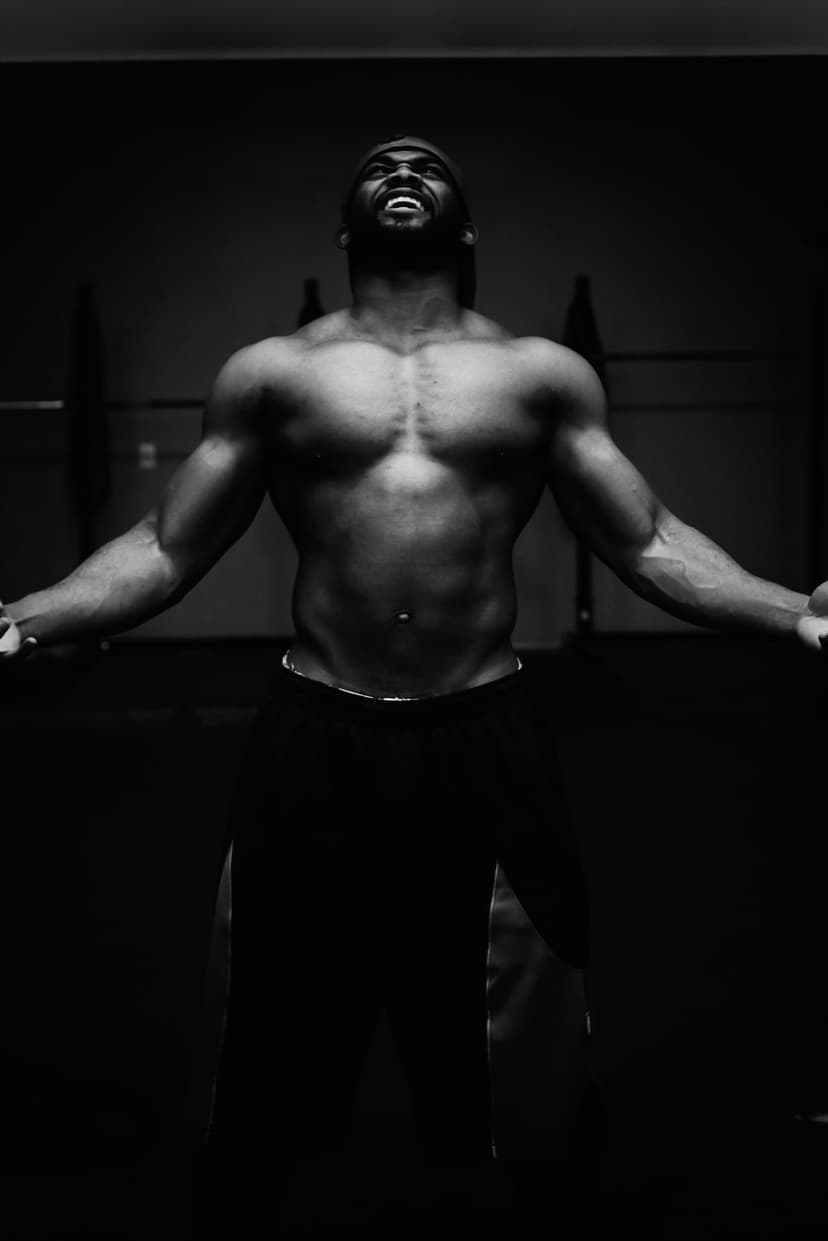 Muscular man with arms outstretched in a dark gym, captured from a low angle with high-contrast monochrome lighting.