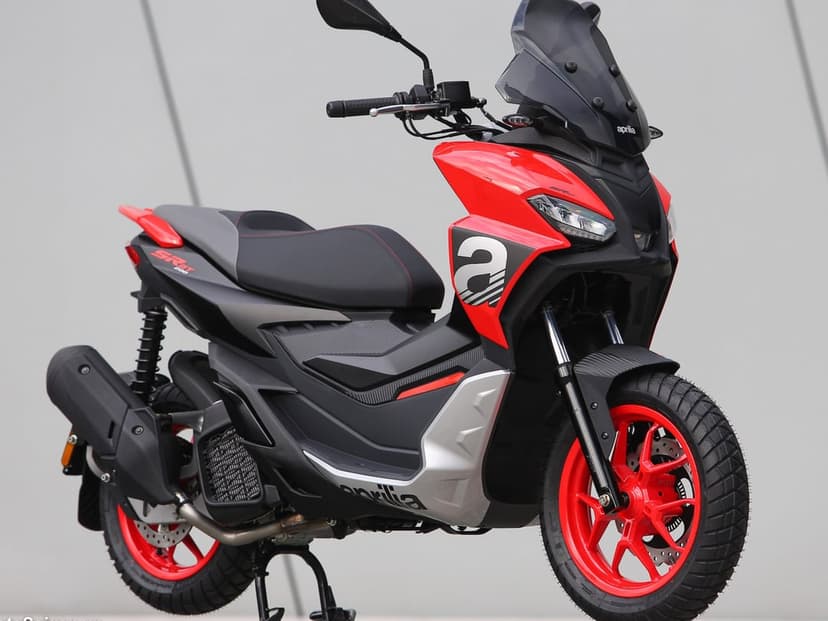 Red and black Aprilia SR GT scooter with red wheels and a tall windscreen on a neutral grey background.