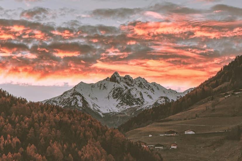 Snow-dusted Dolomite peaks pierce a fiery orange and pink sunset sky above autumn forests and a quiet valley.