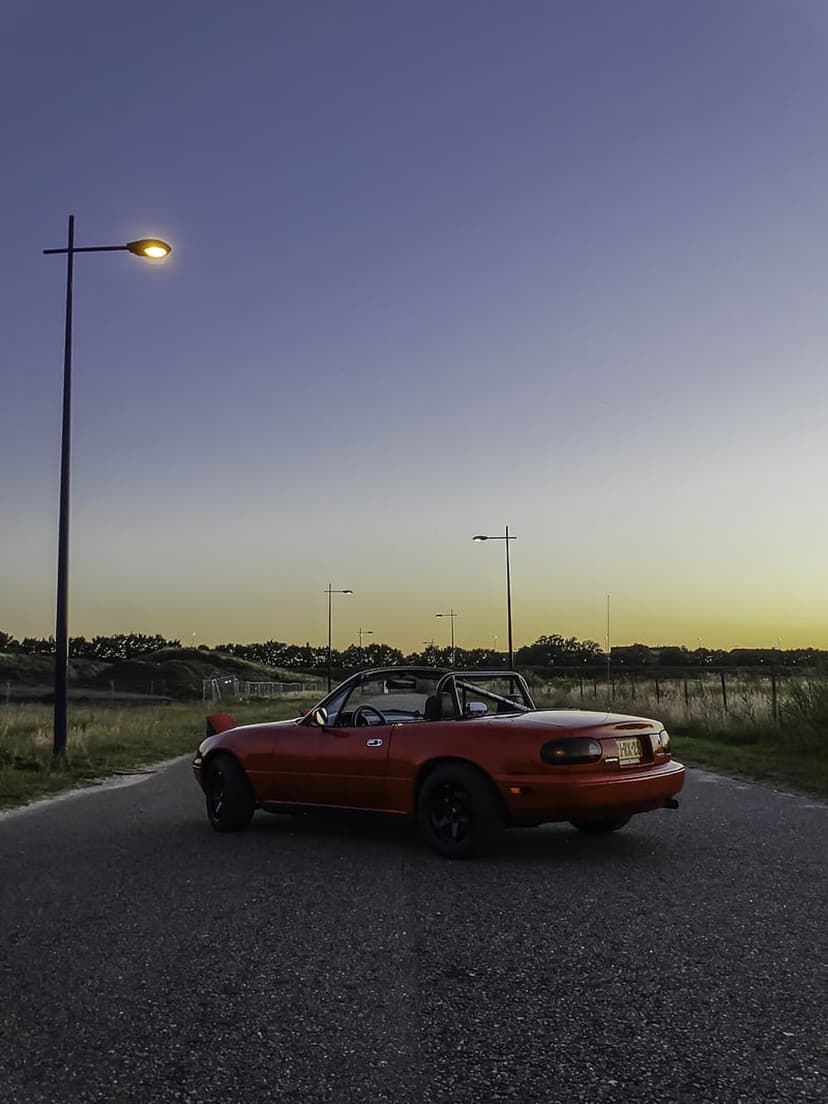 Red Mazda Miata roadster with the top down parked on a road at sunset beneath glowing streetlights during twilight.