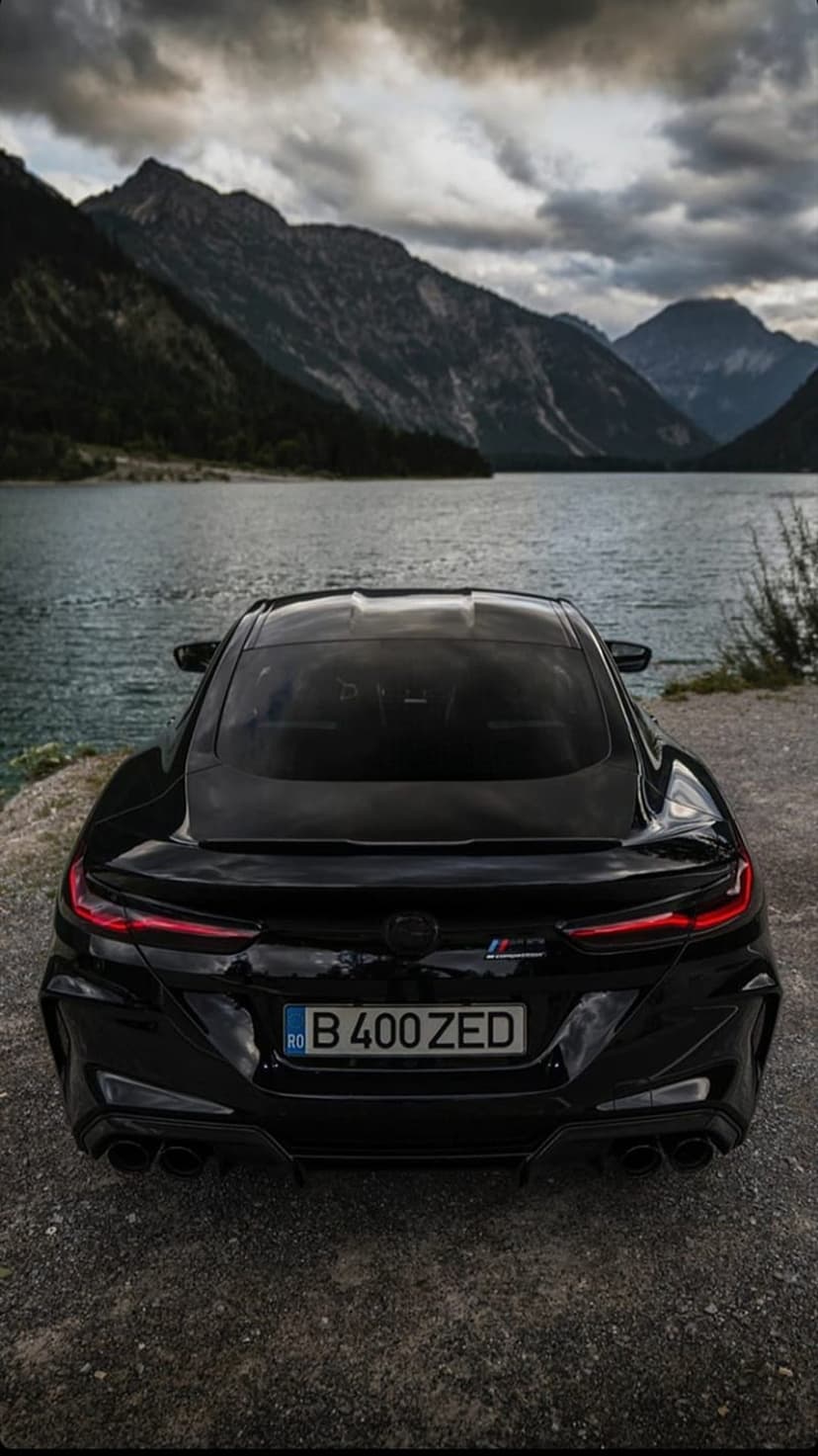 Black BMW M8 Coupe rear view parked by a reflective mountain lake under a cloudy sky during blue hour.