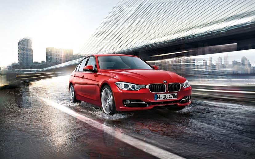 Red BMW sedan driving through a puddle on a wet road under a modern bridge with a city skyline background.
