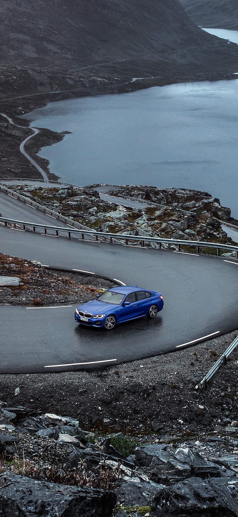 Blue BMW sedan drives on a winding asphalt road alongside a calm lake under a cloudy, grey mountain sky landscape.