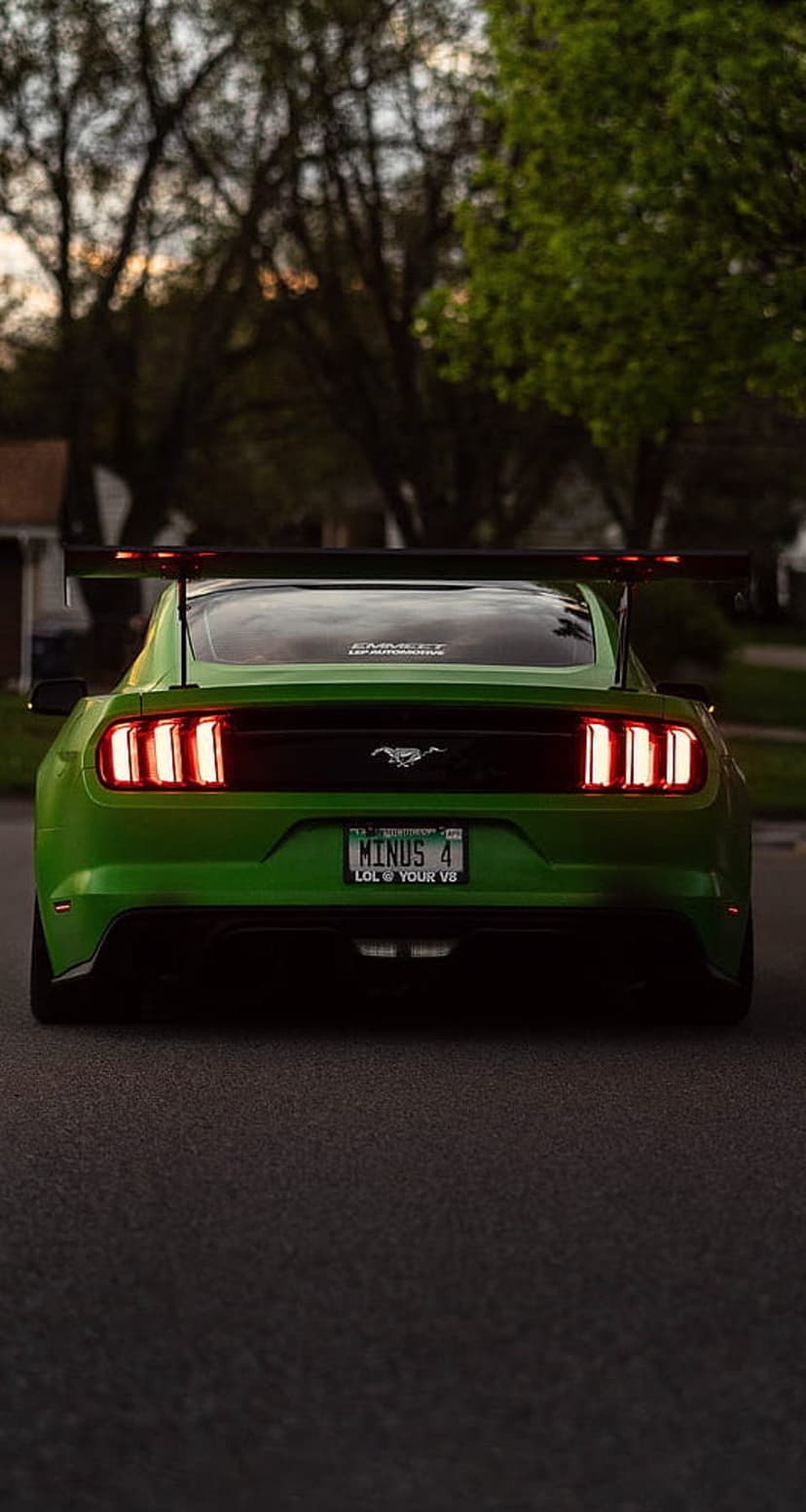 Green Ford Mustang rear view with a massive black spoiler and glowing red taillights on an asphalt road.
