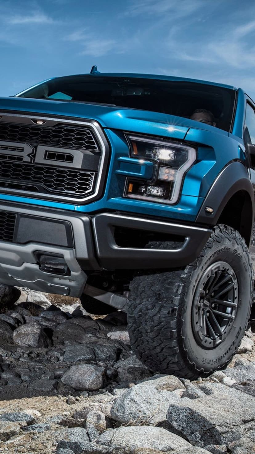 Blue Ford Raptor truck climbing over gray granite rocks with large all-terrain tires and LED headlights.