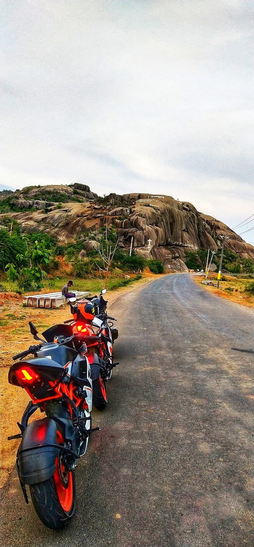 Two orange and black motorcycles park on a paved road beside a steep rocky hill featuring a small white cross.