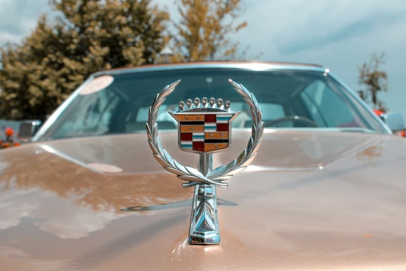 Chrome Cadillac crest and wreath hood ornament mounted on a polished gold vintage car under a cloudy sky.