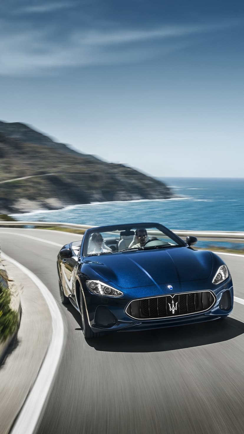 Deep blue Maserati convertible driving on a winding coastal road overlooking the ocean with motion blur.