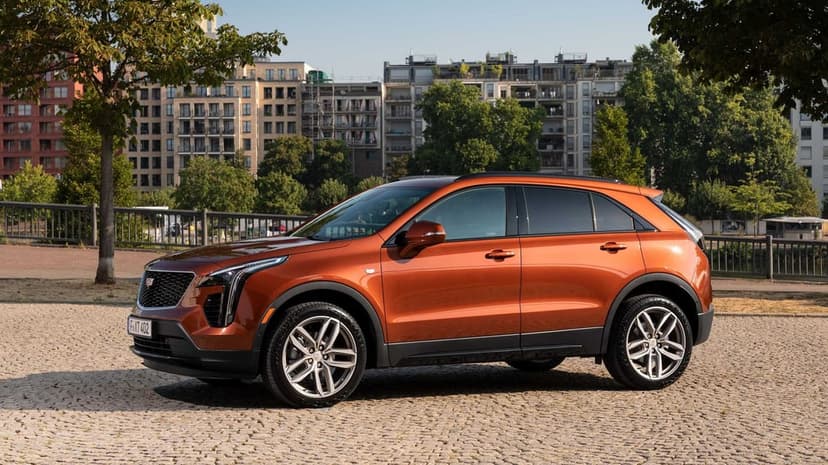 Orange Cadillac XT4 luxury SUV parked on grey cobblestones with city buildings and green trees background.