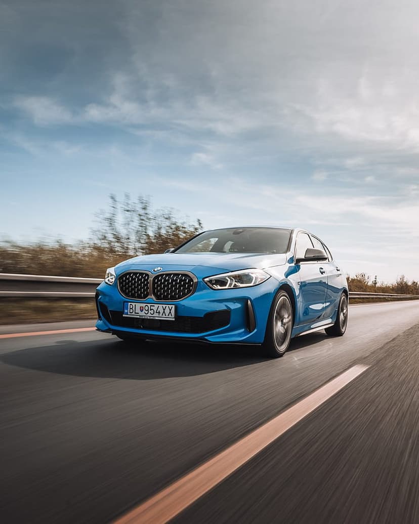 Blue BMW M135i hatchback speeds along a highway with motion blur and sunlight reflecting off the metallic body.