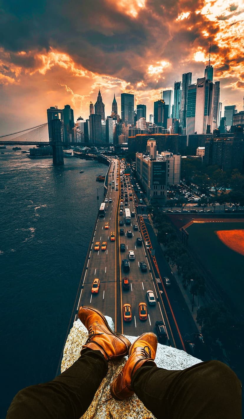 Man's legs in brown boots dangling over a highway and New York City skyline under a fiery sunset sky.