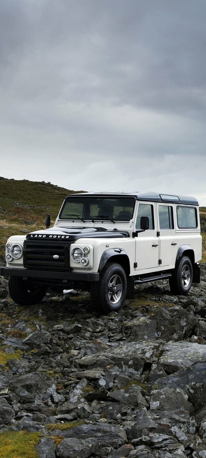 White Land Rover Defender 110 parked on rocky mountain terrain under a gloomy overcast sky.