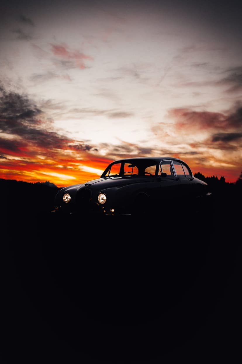 Vintage Jaguar car silhouette against a vibrant orange sunset with illuminated headlights and dramatic clouds.