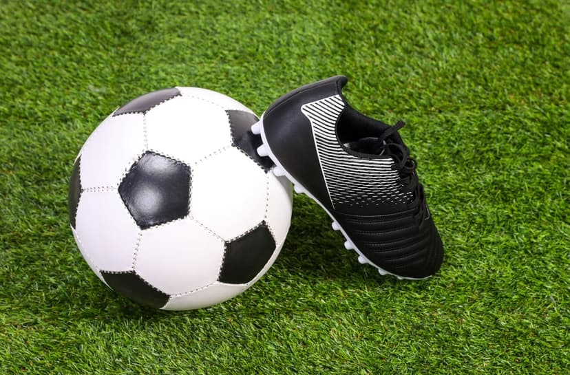 Soccer ball and black cleat with white accents sitting on vibrant green artificial grass under bright lighting.