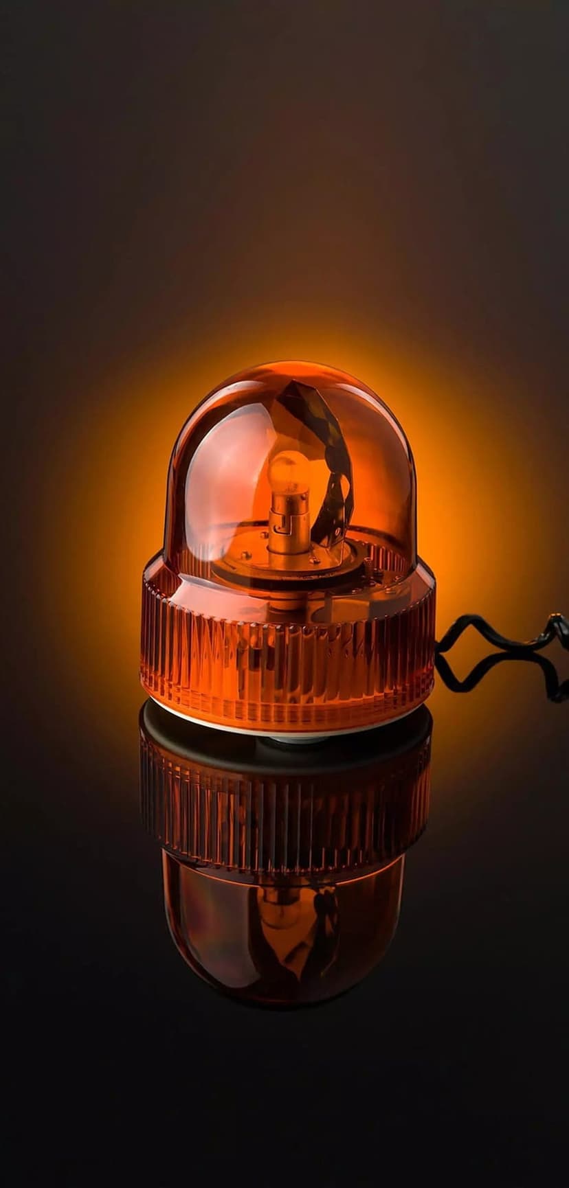 Orange emergency beacon with a ribbed amber dome reflects off a glossy black floor in a dark studio setting.