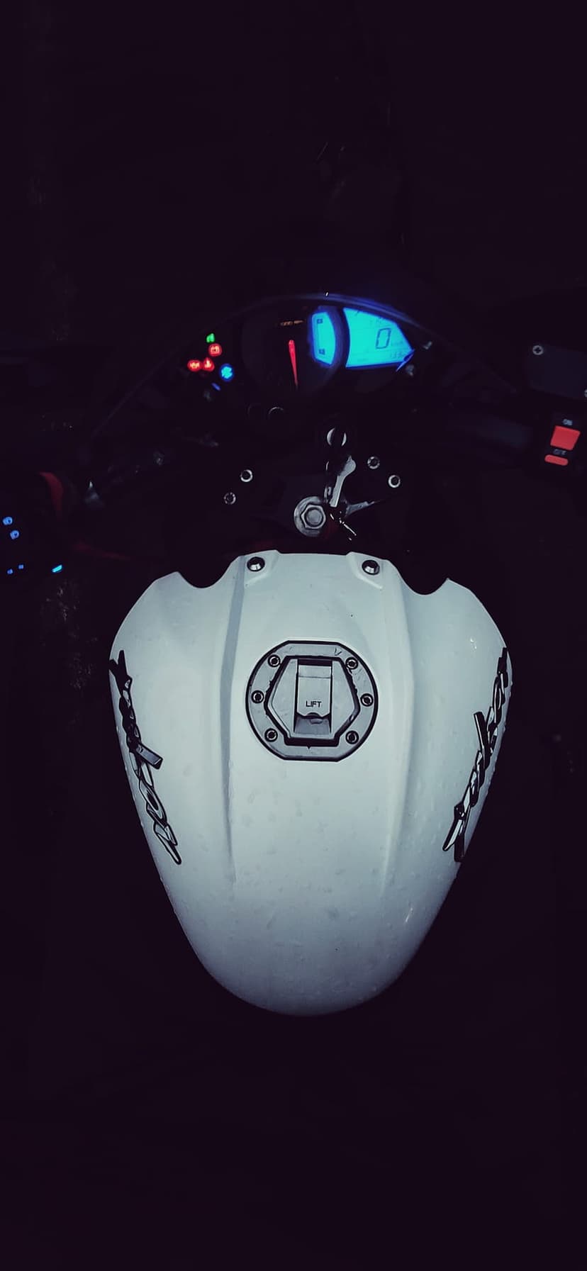 White motorcycle fuel tank and glowing digital dashboard display illuminate a dark nighttime scene from above.