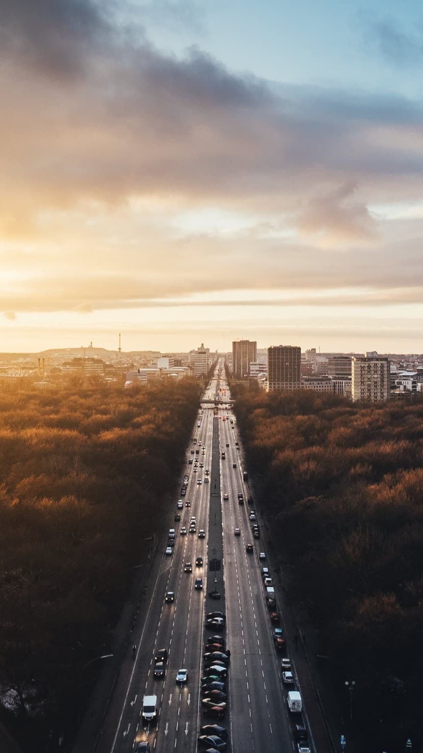 Multi-lane highway with car lights cutting through orange autumn forest toward a hazy sunset city skyline.
