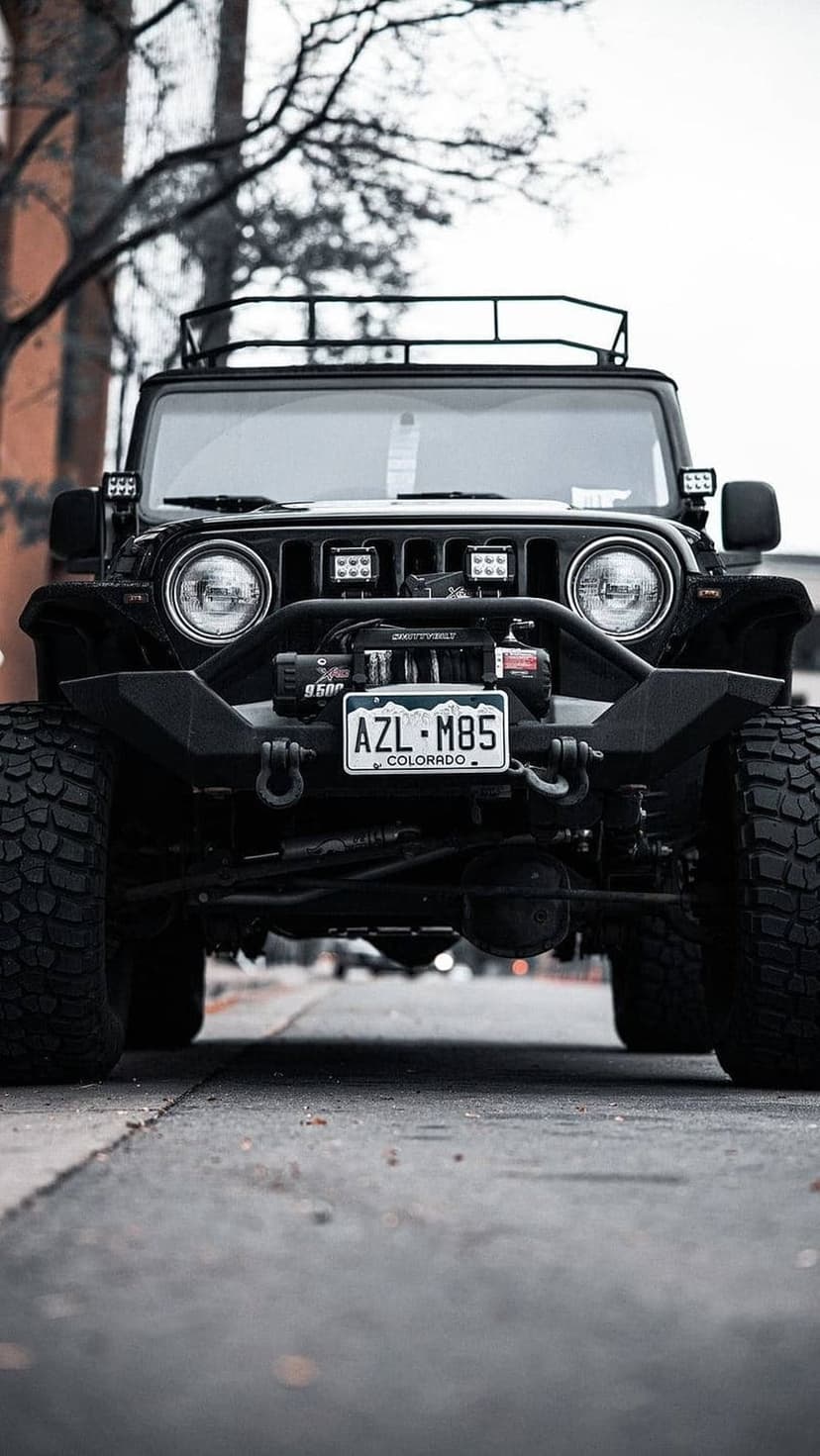 Black Jeep Wrangler with a front bumper winch, roof rack, and Colorado license plate parked on asphalt road.