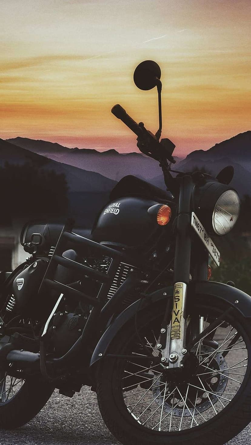 Royal Enfield motorcycle parked on a gravel road with a silhouette of mountains against an orange sunset.